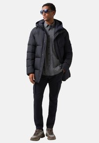 Marineblauwe pufferjack met capuchon, gecombineerd met een geruit overhemd, donkere broek en gestructureerde sneakers. Zonnebril maakt de look af.