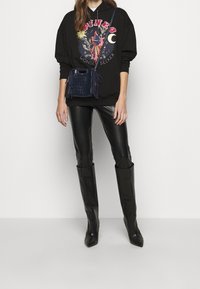 Svart hoodie med grafisk design, glänsande svarta läderbyxor och långa svarta klackstövlar. Matchande marinblå axelväska med fransdetaljer.