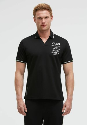 Man wearing black short-sleeve polo with white text and graphics on chest, standing against a plain light background.