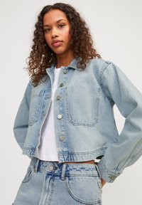 Giacca in denim blu chiaro con design corto, bottoni metallici, due tasche frontali e vestibilità rilassata. Tessuto strutturato con una finitura sbiadita.