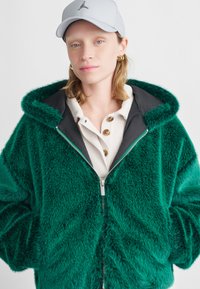 Grüne Fleece-Jacke mit Reißverschluss und schwarzem Innenfutter, ausgestattet mit einer großen Kapuze, kombiniert mit einem cremefarbenen Hemd mit Knöpfen darunter. Texturierte Oberfläche.