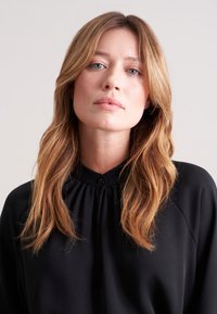 Blusa nera con colletto alto a sbuffo e dettagli con bottoni. Tessuto liscio con una vestibilità comoda e maniche lunghe, che mette in evidenza la texture naturale dei capelli.