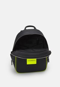 Svart ryggsäck med neongul kant och Emporio Armani-logotyp, framfickan uppdragen som avslöjar svart inredning med zippered ficka.
