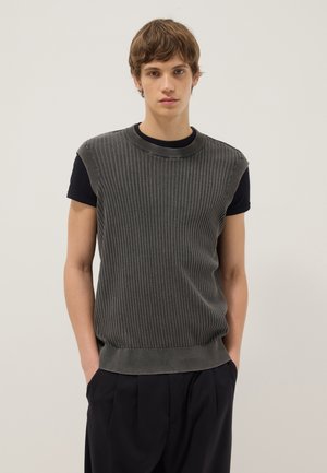 Giovane uomo con capelli castani che indossa un maglione grigio a coste senza maniche sopra una maglietta nera a maniche corte, pantaloni neri, in piedi con le mani nelle tasche.