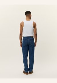 Witte tanktop gedragen met blauwe denim broek, met twee achterzakken en een relaxed fit. De persoon draagt bruine schoenen.