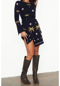 Langärmliges navy Kleid mit goldenen und rosa Palmenstickereien, einem geschichteten Rock und rosa geometrischen Akzenten; kombiniert mit olivgrünen kniehohen Stiefeln.