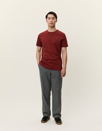 Sarkana kokvilnas T-kreklu ar īsām piedurknēm, apvienota ar pelēkām svītrainām biksēm. Modelis, kas valkā melnās kurpes, stāv uz neitrāla fona.