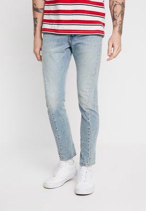Personne portant un jean slim bleu clair, des baskets blanches et une chemise rayée rouge et blanche, montrant des tatouages sur les avant-bras.