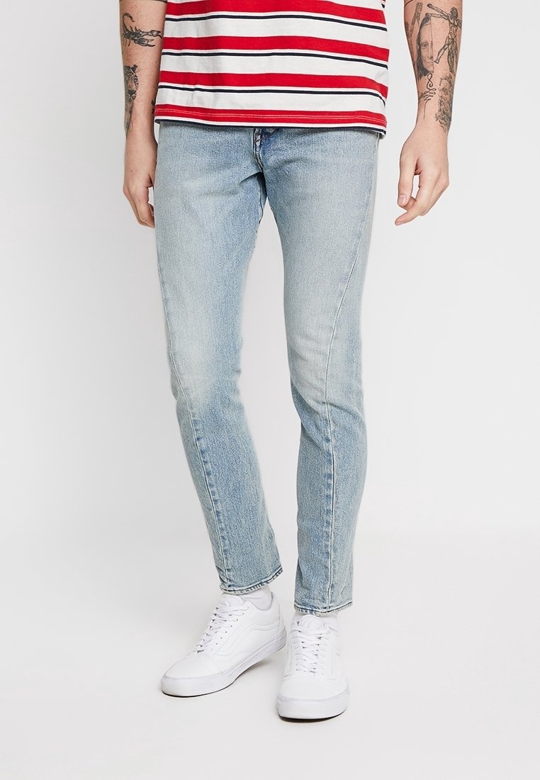 Personne portant un jean slim bleu clair, des baskets blanches et une chemise rayée rouge et blanche, montrant des tatouages sur les avant-bras.