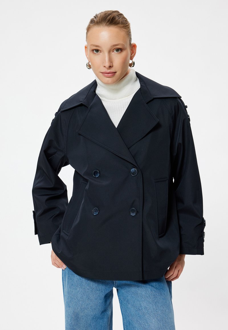 Giacca doppiopetto blu navy con collare a rever, dettagli con bottoni sulle maniche, due tasche e una vestibilità rilassata, abbinata a un dolcevita bianco.
