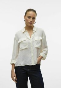 Blusa leggera bianca con chiusura a bottoni sul davanti, due tasche sul petto e maniche arrotolate; indossata con jeans scuri. Texture liscia.