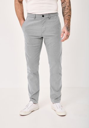 Man wearing light gray slim-fit pants with pockets, a white shirt, white sneakers, and visible arm tattoos on a plain background.