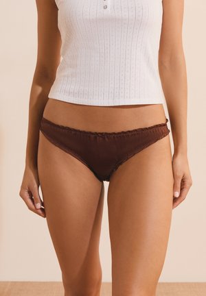Mujer con ropa interior estilo bikini de satén marrón oscuro y una camiseta sin mangas blanca de canalé, de pie frente a un fondo beige liso.