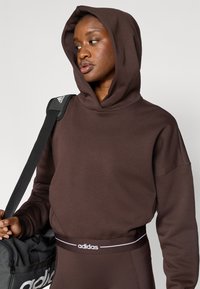 Tumepruun kapuutsiga pusa pehmest materjalist, millel on oversized-lõige ja valge adidas logo. Mudel hoiab musta spordikotti.