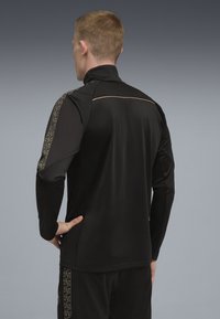 Svart atletisk jacka med hög krage, med strukturerat svart tyg och ett mönstrat guldaccent längs axeln.