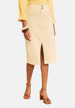 Kvinde iført en beige knælængde nederdel med frontslids, gul langærmet bluse, brune hælede sandaler og chunky armbånd på venstre håndled.