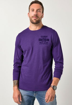 Lila langärmliges T-Shirt mit Rundhalsausschnitt, ausgestattet mit einem schwarzen Druck "Canoe Southern Ireland" auf der linken Brust. Weicher Stoff.