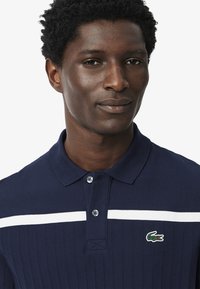 Polo náutico con un patrón texturizado, que presenta una raya horizontal blanca y un pequeño logo de cocodrilo verde en el lado izquierdo del pecho.