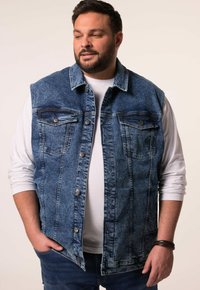 Denimová bezrukávová bunda se dv ěma kapsami na prsou, světle modrá barva, kovové knoflíky a kontrastní švy. Nositá přes bílé tričko s dlouhým rukávem.