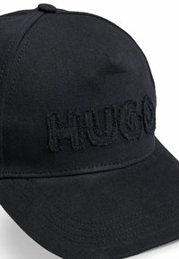 Svart keps av twilltyg med en böjd skärm och broderad "HUGO"-logotyp i en sliten stil.