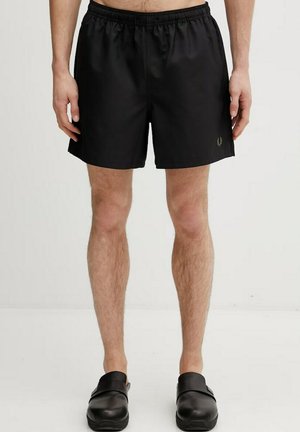 Uomo che indossa pantaloncini con vita elastica neri e scarpe slip-on nere, in piedi contro uno sfondo chiaro semplice.