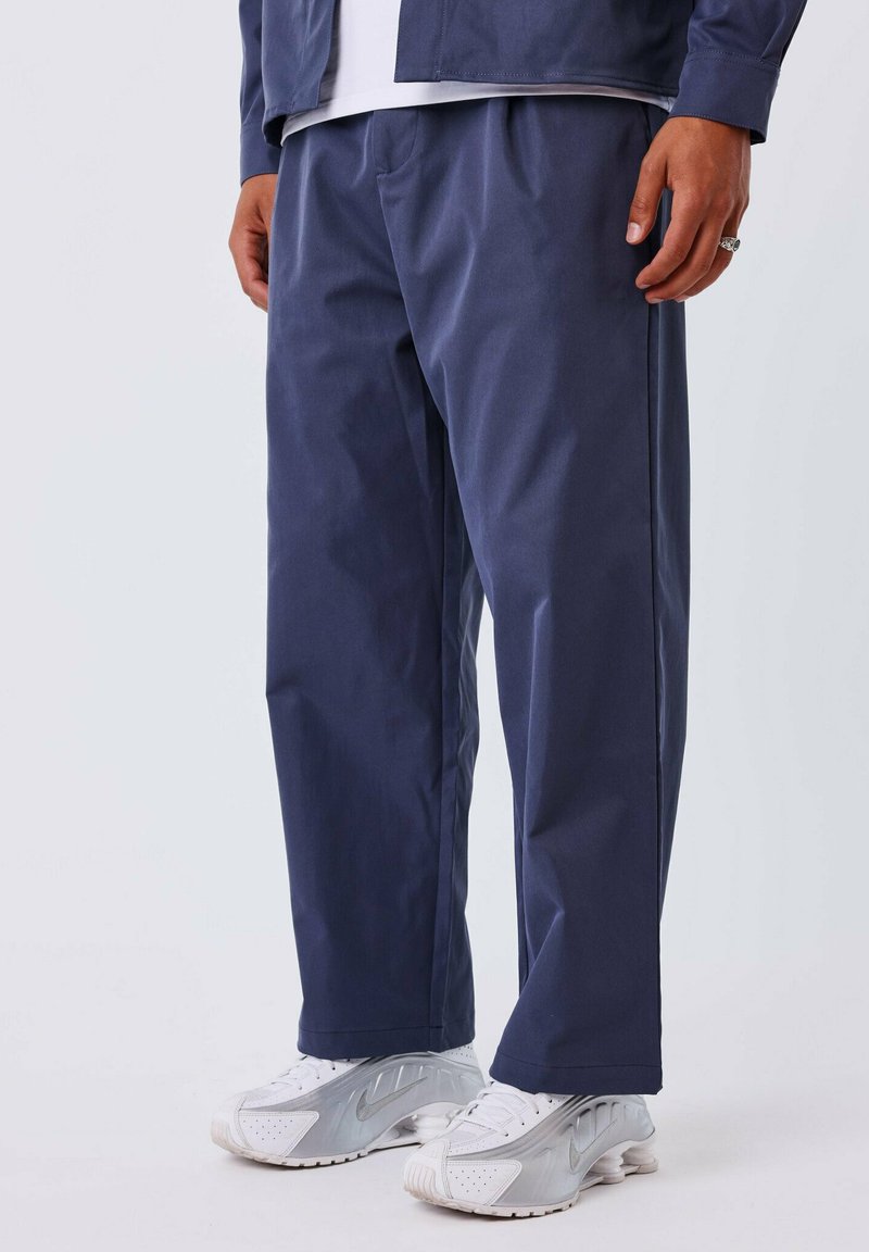 Marineblauwe, losse broek van een glad materiaal met zijzakken. Gekleed met witte sportieve schoenen met zilveren accenten.