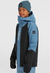 Blauw en zwart geïsoleerd jack met een hoge kraag, ritsluiting en logodetail. Inclusief zwarte handschoenen en een blauwe gebreide muts.