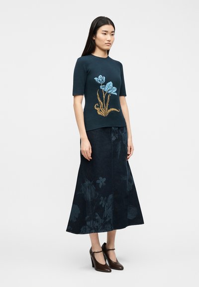 Γυναίκα που φοράει μπλε σκούρο κοντομάνικο τοπ με μπλε και καφέ ανθισμένο σχέδιο, midi φούστα με μπλε ανθικό μοτίβο και καφέ ψηλοτάκουνα.