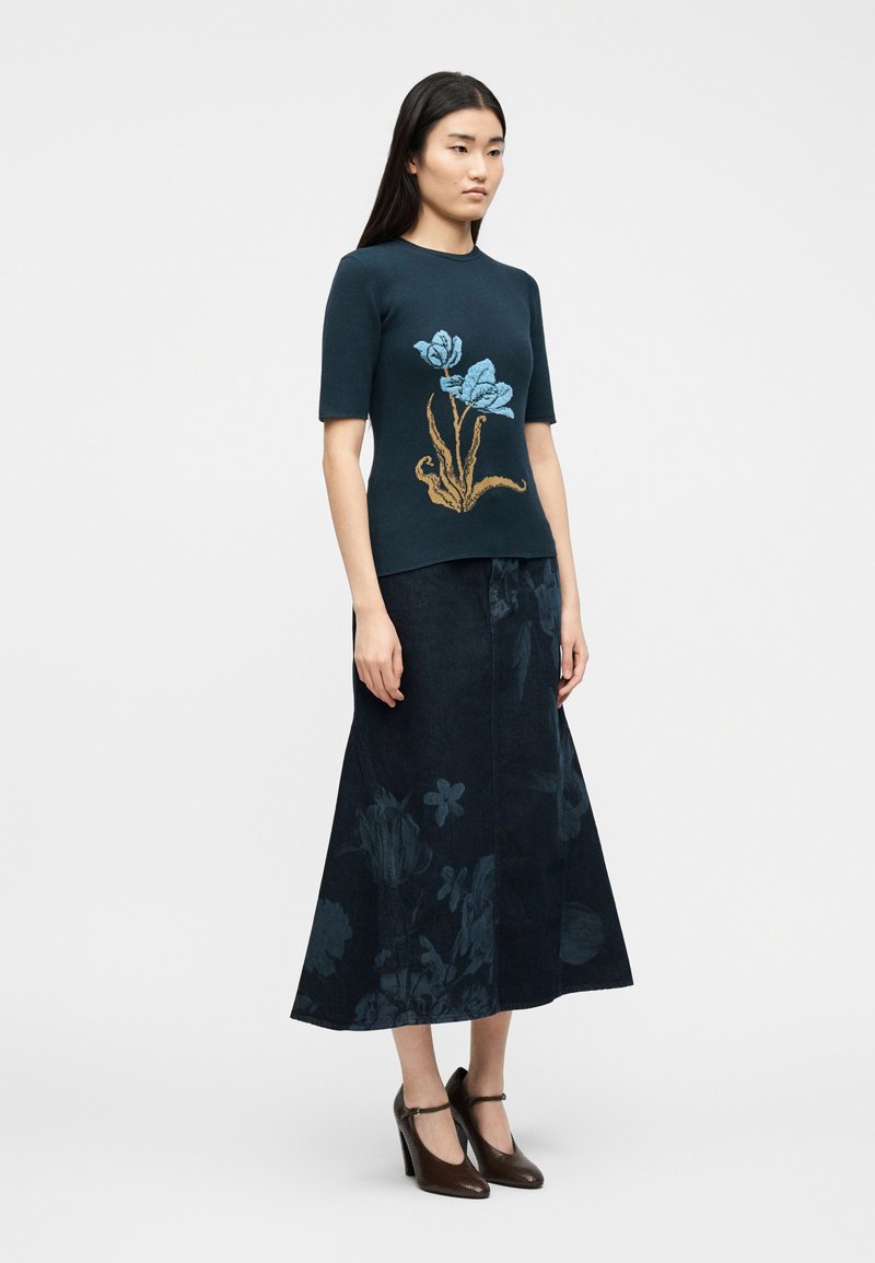 Donna che indossa una maglietta a maniche corte blu scuro con un motivo floreale blu e marrone, gonna midi a fantasia floreale blu scuro e tacchi marroni.