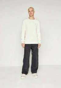 White long-sleeve shirt with a round neck, paired with loose-fitting black pants featuring zippered pockets and white sneakers.