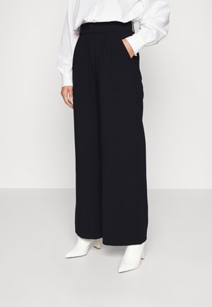 Kobieta w wysokich, szerokich czarnych spodniach, białej, długorękawowej bluzce i białych botkach z ostrym noskiem, stojąca z jedną ręką w kieszeni.