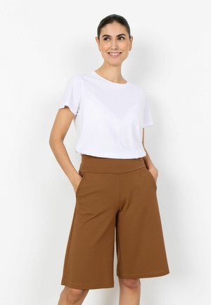 Usmievavá žena s tmavými vlasmi v copu, oblečená v bielom tričku s krátkym rukávom a hnedých širokých nohaviciach po kolená s vysokým pásom, ruky v vreckách.