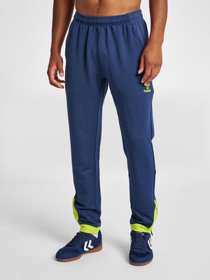 Mand iført marineblå træningsbukser med gule accenter og marineblå sportssko med hvide chevron-logoer.