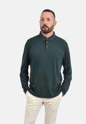 Uomo con la barba che indossa una polo verde scuro a maniche lunghe e pantaloni chiari, in piedi con le mani nelle tasche, guardando di lato.