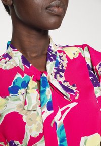 Květinová blůza s růžovým pozadím, s krčním vázáním, živé barvy včetně modré, zelené a béžové, s hedvábnou texturou.