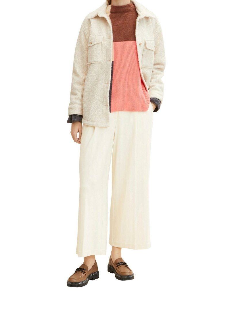 Jasna, teksturowana kurtka z dwoma kieszeniami na piersi, noszona na swetrze z blokami kolorów (czerwony, różowy, brązowy) oraz szerokich, kość słoniową spodni. Brązowe mokasyny.