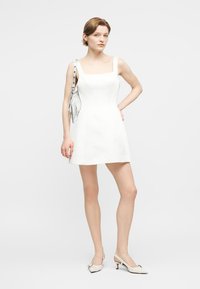 Kvinde står iført en kort hvid ærmeløs kjole, hvide flade sko og en sølv skuldertaske.