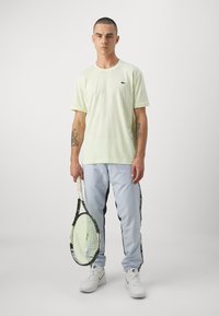 Lys grøn kortærmet t-shirt og lys blå bukser med sorte sideliner, holder en tennisracket; hvide sportssko.