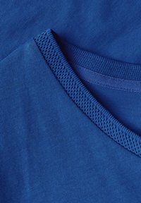 Blå tyg t-shirt med en slät yta. Har en ribbad rund halsringning och ett lager på lager krage design, vilket visar sömnadsdetaljer.