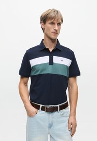 Laivaston polo-paita, jossa valkoiset ja turkoosit vaakasuorat raidat, lyhyet hihat, kaulukset ja logotyyppinen yksityiskohta. Yhdistetty vaaleansinsisiin farkkuihin ja ruskeaan vyöhön.