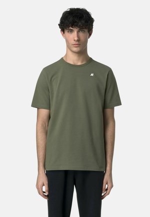 Giovane uomo con capelli ricci e scuri che indossa una t-shirt verde oliva a maniche corte con un piccolo logo bianco sul petto, in piedi davanti a uno sfondo semplice.