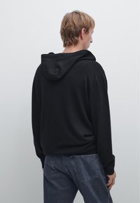 Schwarzer oversized Hoodie mit Kordelzugkapuze, langen Ärmeln und lässiger Passform, kombiniert mit dunkelblauen Jeans, von hinten sichtbar.