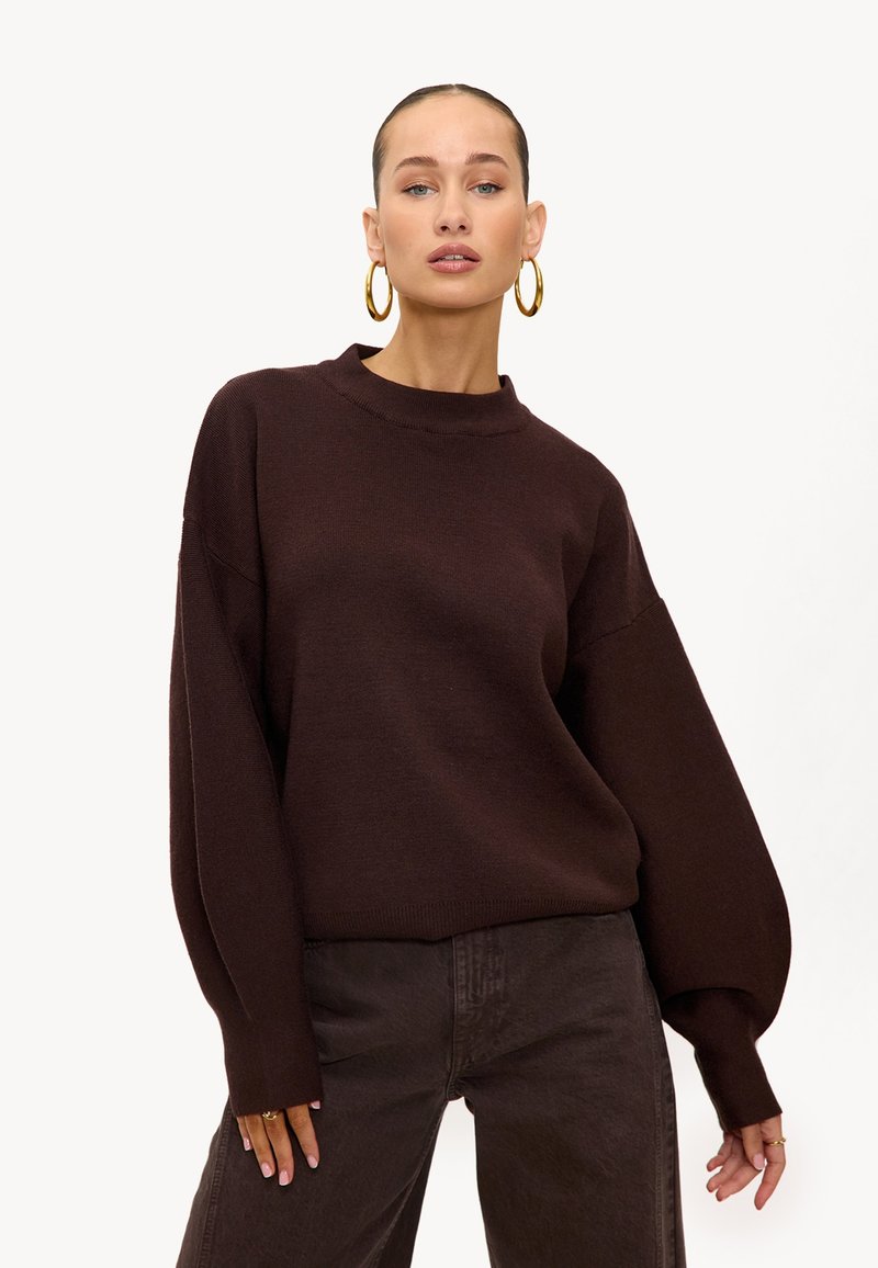 Pull marron en maille avec un col haut et des manches surdimensionnées, associé à un pantalon marron foncé ; design minimaliste et texture douce.