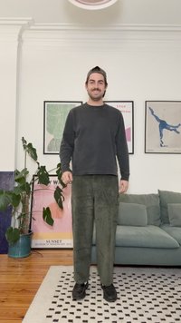 Zwart shirt met lange mouwen, olijfgroene corduroy broek en zwarte sneakers. Staand op een patroonmat in een woonkamer met kunst aan de muren.