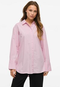Šviesiai rožinė marškinė su apykakle, ilgomis rankovėmis ir plačiomis apykaklėmis, pagaminti iš glotnaus medvilnės audinio. Esminė detalė: priekinis sagčių užsegimas.