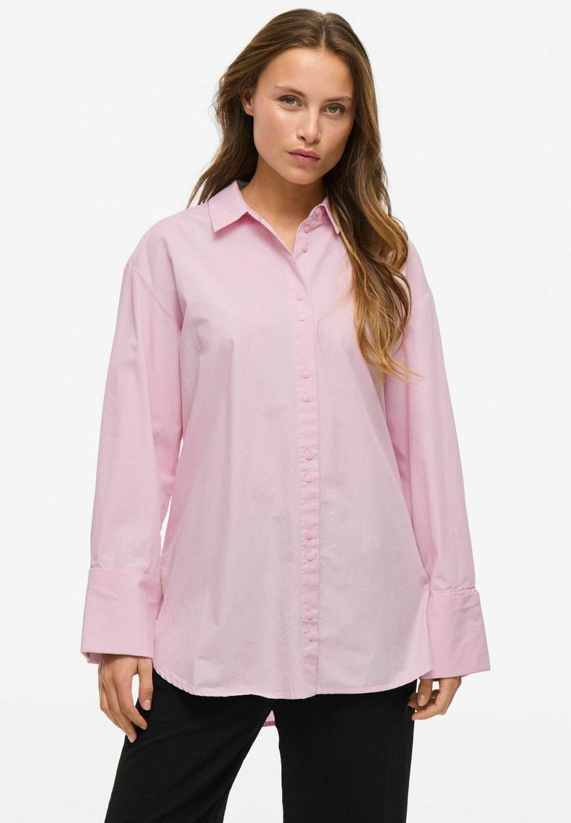 Šviesiai rožinė marškinė su apykakle, ilgomis rankovėmis ir plačiomis apykaklėmis, pagaminti iš glotnaus medvilnės audinio. Esminė detalė: priekinis sagčių užsegimas.