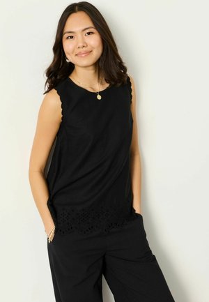 Young woman with long dark hair wears sleeveless black top with lace hem and black pants, standing with hands in pockets, smiling gently.