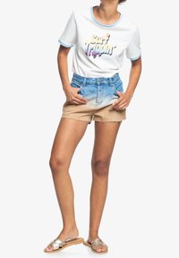 Camiseta blanca de manga corta con ribete azul y gráfico de "Surf Trippin'". Pantalones cortos de mezclilla en azul claro a beige con dobladillo deshilachado. Chanclas beige.