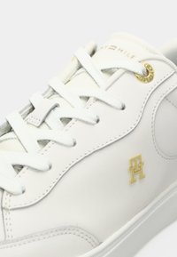 Hvite lær sneakers med avrundet tå, grå sømmer, flate hvite snører og et gulllogo på siden. Glatt tekstur.