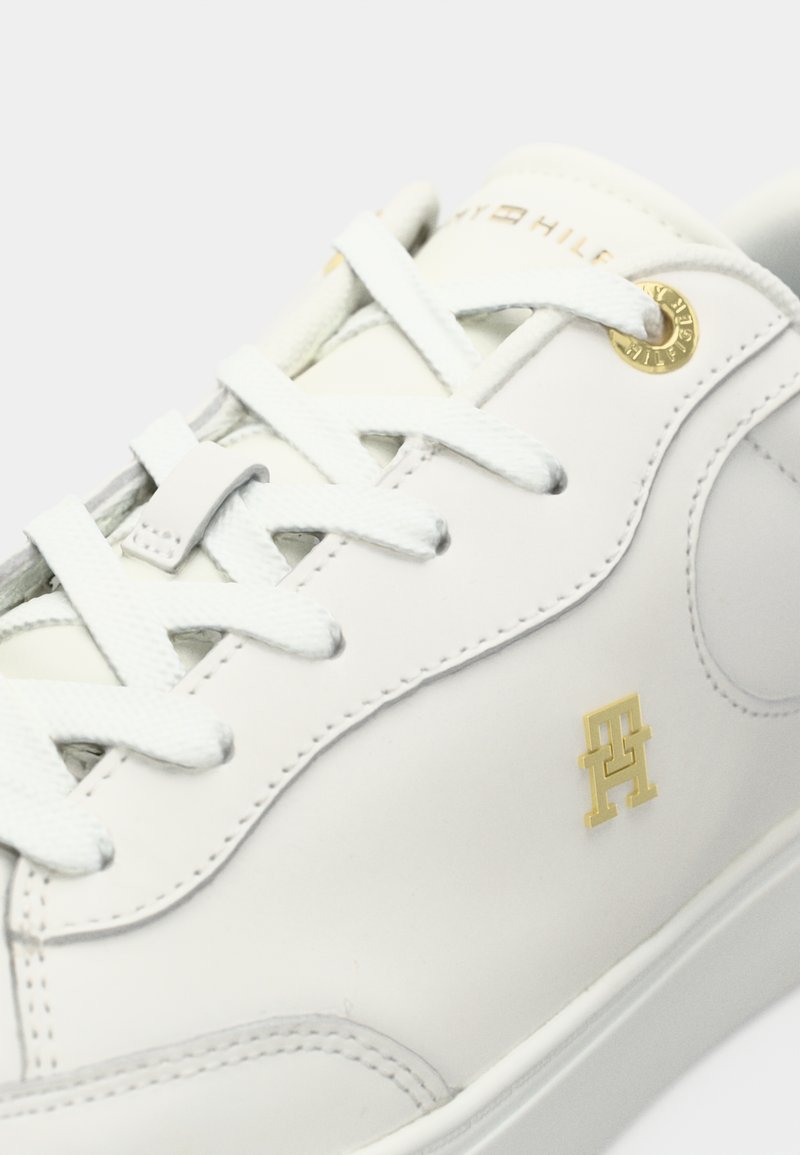 Weiße Ledersneaker mit abgerundeter Zehenpartie, grauen Nähten, flachen weißen Schnürsenkeln und einem goldenen Logodetail an der Seite. Glatte Textur.